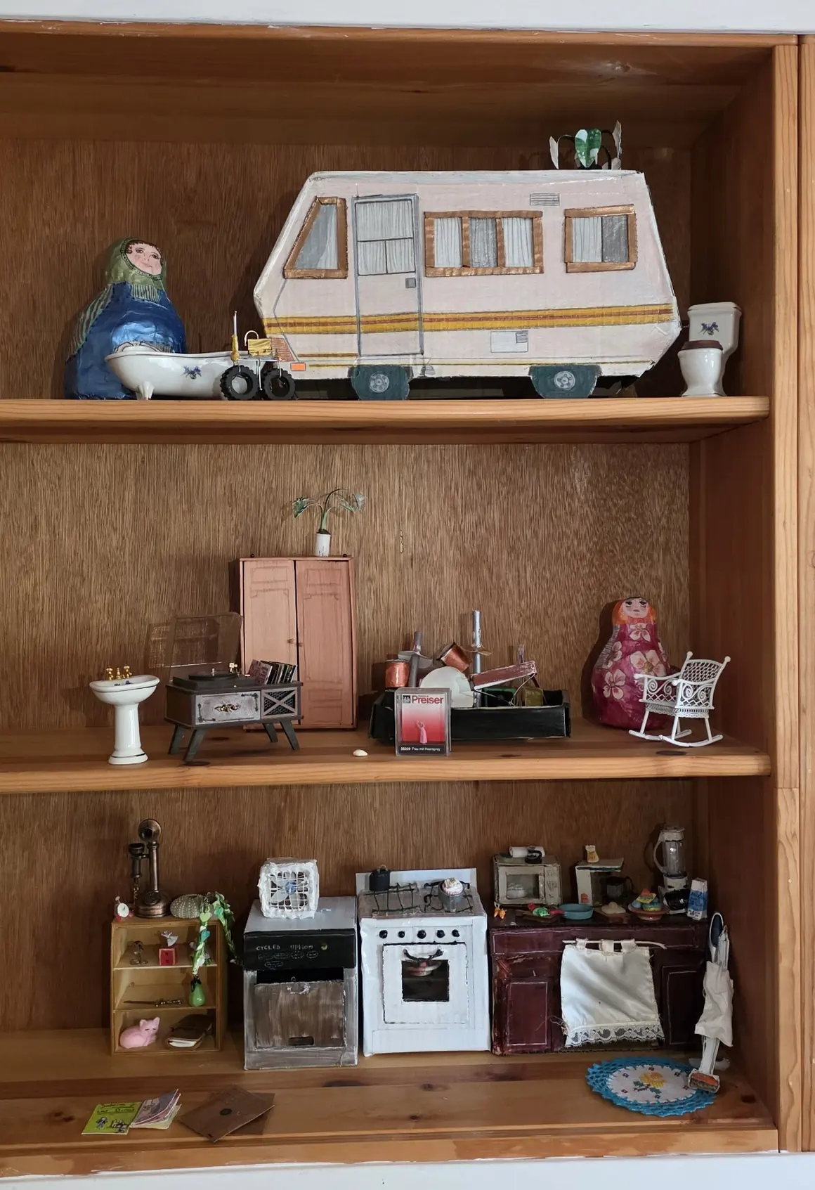 Estante de madera con tres baldas llenas de miniaturas: una caravana de papier mâché arriba, un tocadiscos y una matrioska en el medio, y una cocina en miniatura abajo.